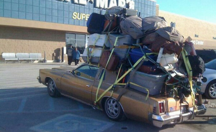 Old car parked outside Walmart, overloaded with numerous bags and items secured by straps on its roof and bed, Walmart people.