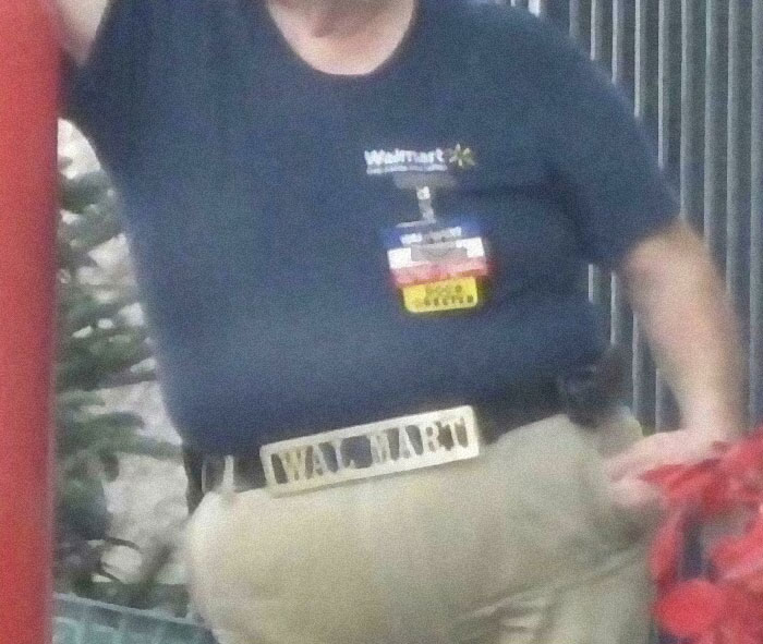 Walmart people employee wearing a navy shirt with Walmart logos and a large Walmart belt buckle indoors.