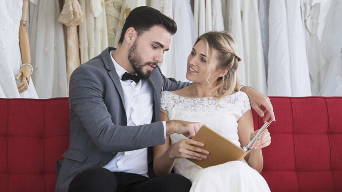 Groom and fiancée in wedding attire discussing finances and debt issues on a red couch before wedding planning. Groom and fiancée in wedding attire discussing finances and debt issues on a red couch before wedding planning.