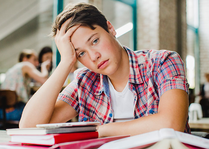 Joven con expresión pensativa y celos, sentado en mesa con libros abiertos, reflexionando sobre consecuencias inesperadas.