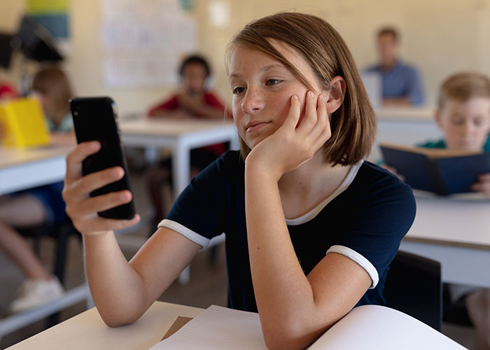 Niña sentada en clase mirando su teléfono móvil, con expresión pensativa sobre historias de consecuencias no intencionadas.