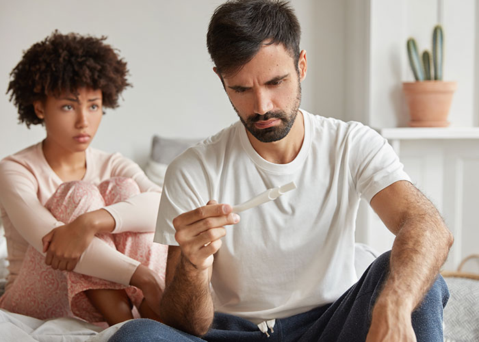 Hombre preocupado mirando prueba de embarazo mientras pareja en segundo plano refleja celos e incertidumbre emocional.
