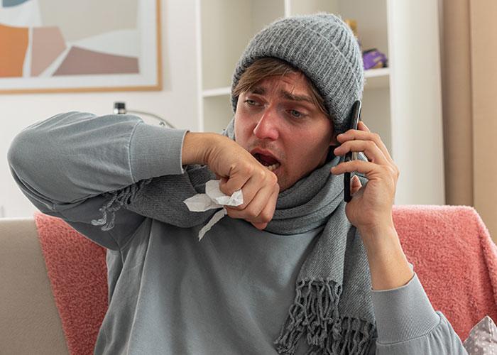Hombre con gorro y bufanda hablando por teléfono, tosiendo, mostrando síntomas de resfriado y consecuencias imprevistas.