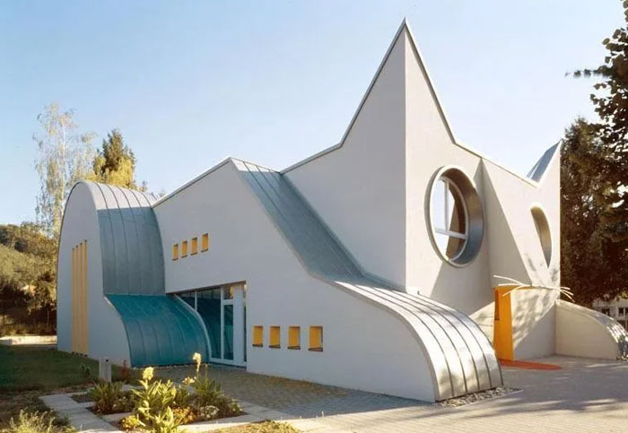 Unique bread-shaped bread factory building with curved and angular design elements surrounded by trees and greenery.