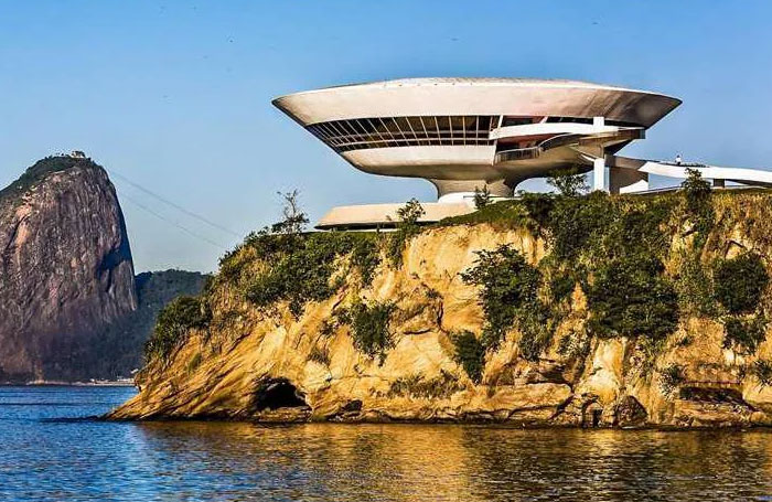 Futuristic bread-shaped bread factory building perched on a rocky coastline with clear blue sky and water.
