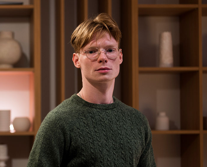 Young man wearing glasses and green sweater posing indoors, related to DNA tests revealing shocking family information.