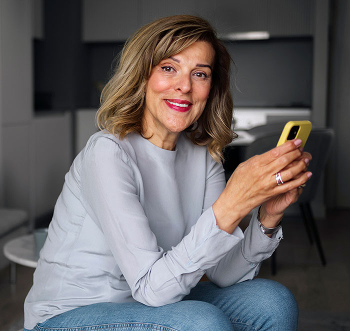 Middle-aged woman holding a yellow phone, smiling and sitting indoors, relating to DNA tests and family relationships.
