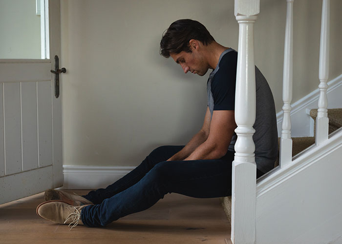 Man sitting on floor near stairs, looking down sadly, representing husband&rsquo;s ruined reputation after wife leaves him.