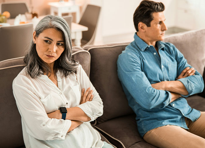 Middle-aged couple sitting apart on couch with arms crossed, depicting dad calling out wife over daughter treating boyfriend like ATM. Middle-aged couple sitting apart on couch with arms crossed, depicting dad calling out wife over daughter treating boyfriend like ATM.