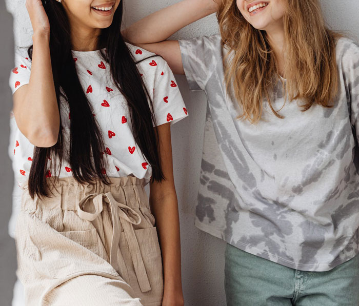 Two teenage girls smiling and talking, one wearing a heart print shirt, illustrating woman helps detangle daughter's friend&rsquo;s hair.