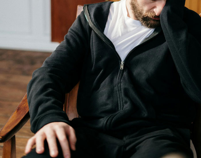 Man in black hoodie sitting with head in hand looking stressed or upset indoors on wooden floor background