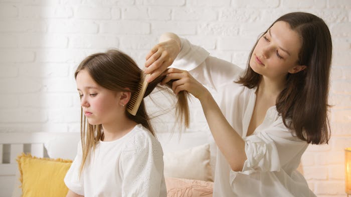 Woman gently helps detangle daughter&rsquo;s friend&rsquo;s hair in a calm home setting, focusing on care and patience.