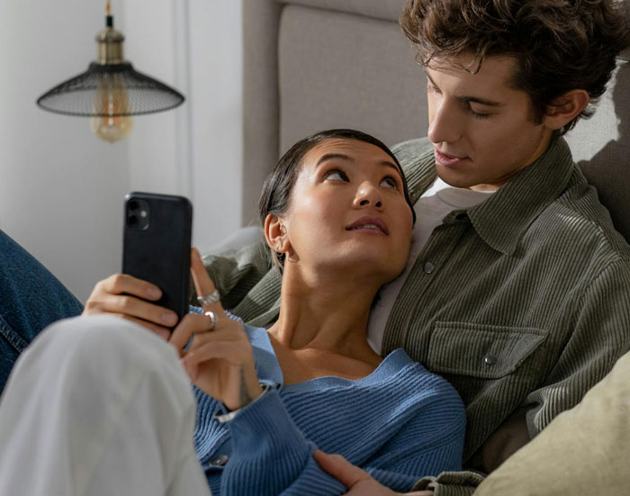 Woman showing proof of cheating on phone to her husband while sitting closely on couch at home.