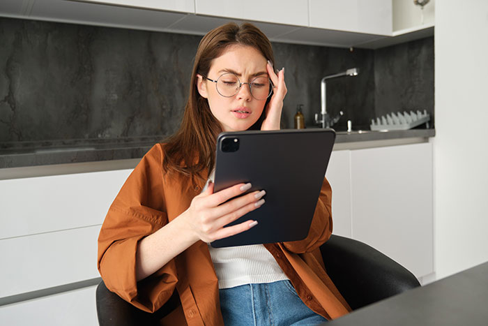 Young woman in glasses reading tablet with a worried expression after finding secret texts about her appearance.