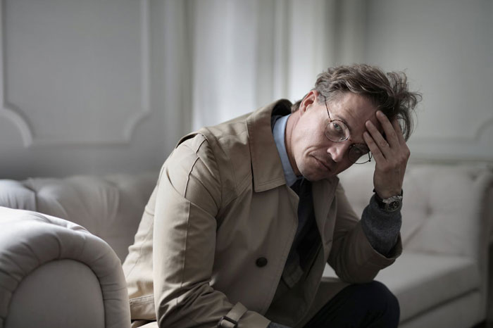 Man in beige trench coat and glasses sitting on couch, looking distressed and unsure about affair partner's baby situation.