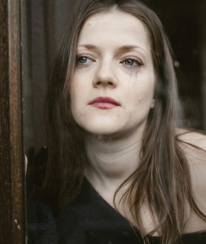Woman with smudged makeup looking out a window, reflecting on uncertain affair partner's baby and complex family ties.