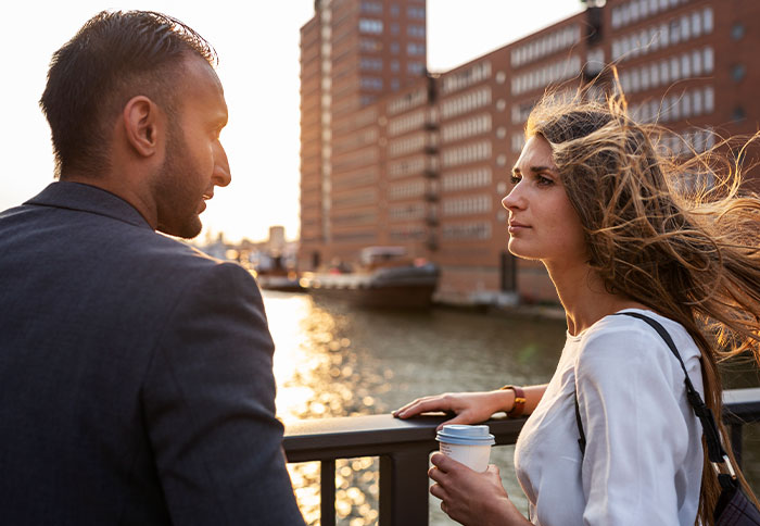 Woman getting a reality check after regretting a divorce, talking seriously with a man near waterfront buildings at sunset.
