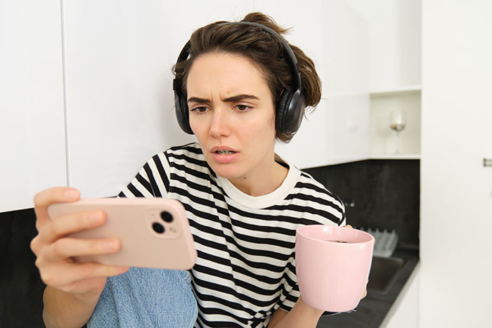 Young woman with headphones holds phone and pink mug, looking shocked while reading social media about coworker conflict. Young woman with headphones holds phone and pink mug, looking shocked while reading social media about coworker conflict.