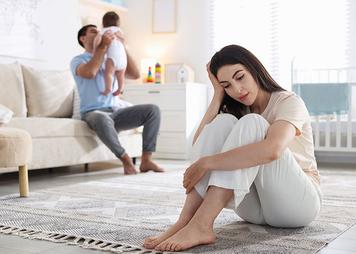 Tired mom sitting on floor looking stressed while father plays with baby in living room during family time.