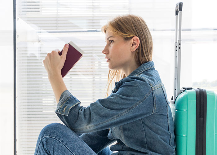 Woman in denim outfit sitting with suitcase, holding passport and looking away, depicting a mom running away from her kids.