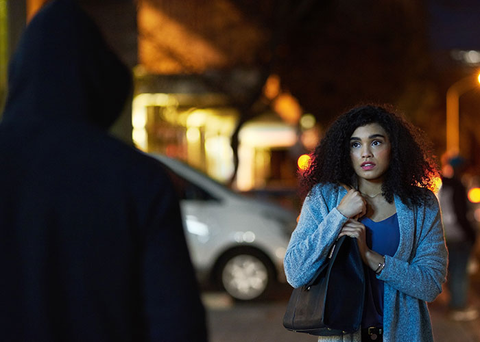 Woman standing on a dimly lit street looking concerned as a man in a hoodie approaches, highlighting stalker fear period pad.