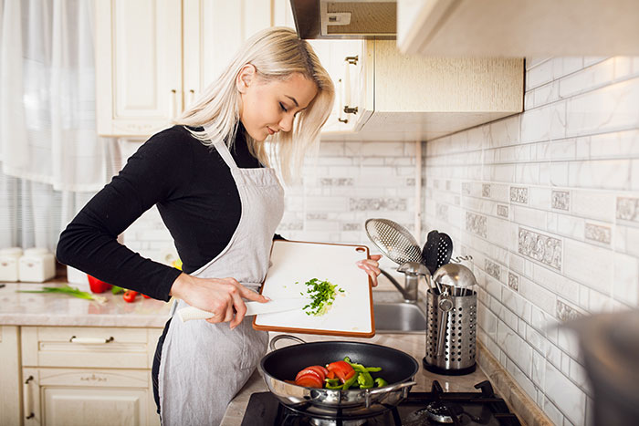 Young woman in apron cooking vegetables in kitchen, illustrating traditional wives concept in a modern setting.
