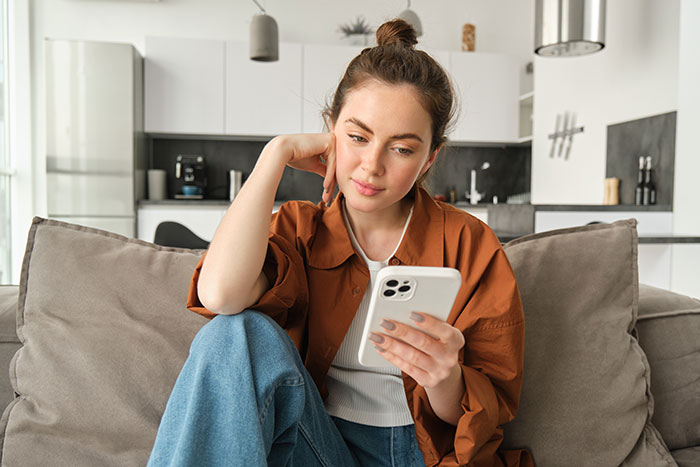 Woman in casual outfit sitting on couch looking thoughtfully at smartphone, reflecting on wedding photo controversy.