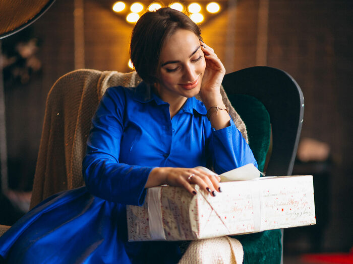 Woman in blue dress sitting in cozy armchair, holding holiday gift that might reveal painful truth about relationship.