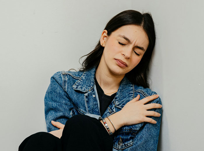 Woman in denim jacket sitting against a wall looking upset, illustrating a woman blinded by flowers from her son.