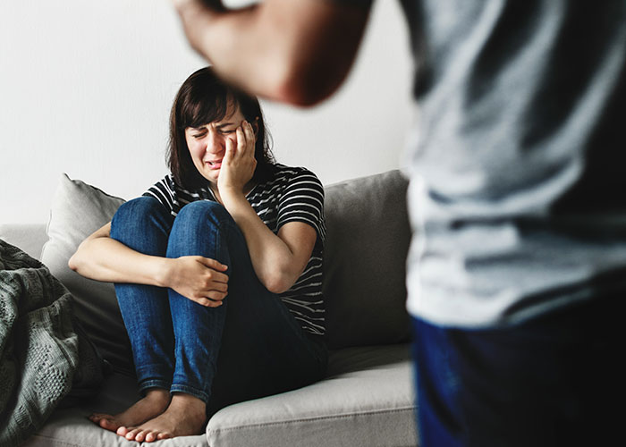 A woman sits crying on a couch, upset, while a person stands in the foreground, symbolizing an insensitive gym membership gift.