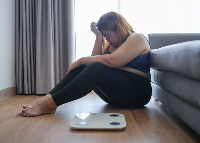 Woman sitting on the floor next to a scale, looking upset and overwhelmed by a gym membership gift.