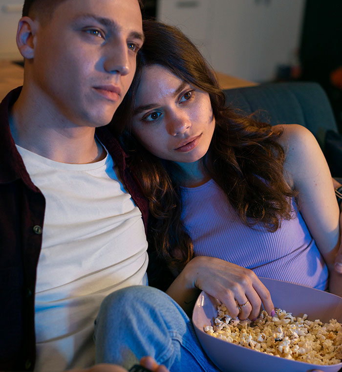 Couple sitting closely on a couch with popcorn, looking pensive in a low-lit room after a proposal story discussion.