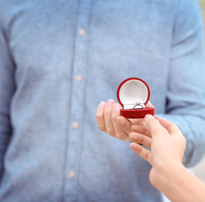 Person in blue shirt holding an open ring box with an engagement ring during a proposal moment.