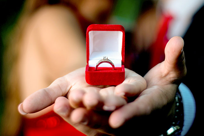 A close-up of a hand holding an open red velvet ring box with a diamond engagement ring inside.
