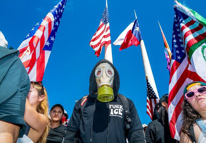 Person wearing a gas mask surrounded by protesters holding American and Texas flags in vibrant street photography by Perry Hall.