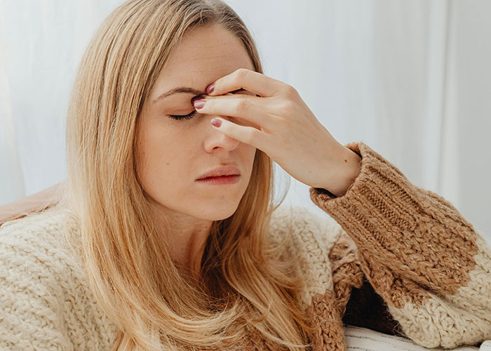 Woman in cozy sweater with eyes closed, pinching bridge of nose in frustration over baby shower conflict.