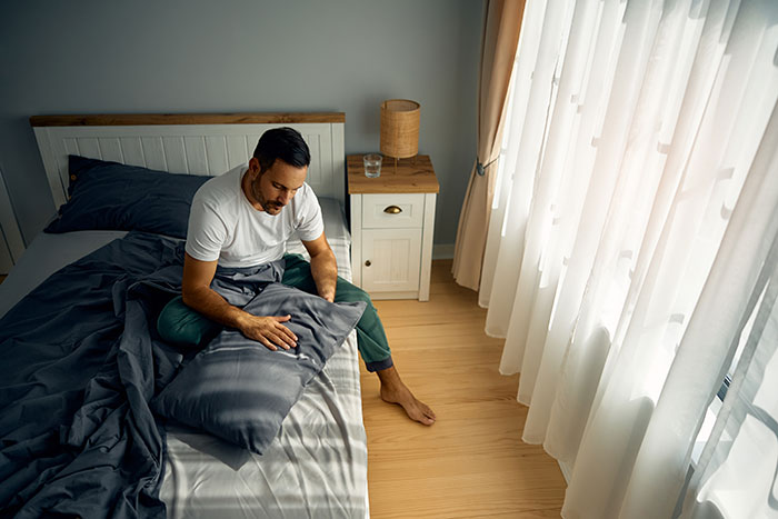 Man sitting on bed holding a pillow, appearing distressed in a bright room with sunlight through sheer curtains.