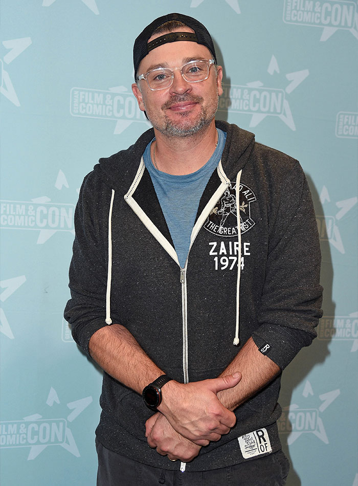 Man at a fan convention wearing glasses, a black cap, and a dark hoodie, a 2000s teen heartthrob now aged 48.