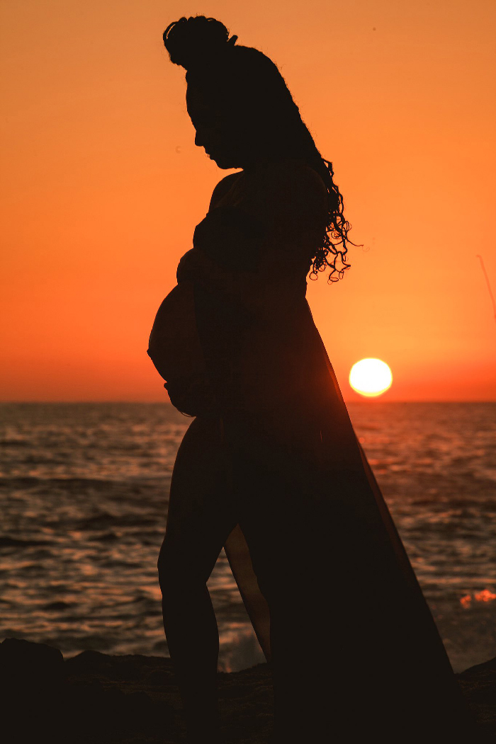 Silhouette of pregnant woman on a beach at sunset holding belly, creative maternity photoshoot scene with orange sky