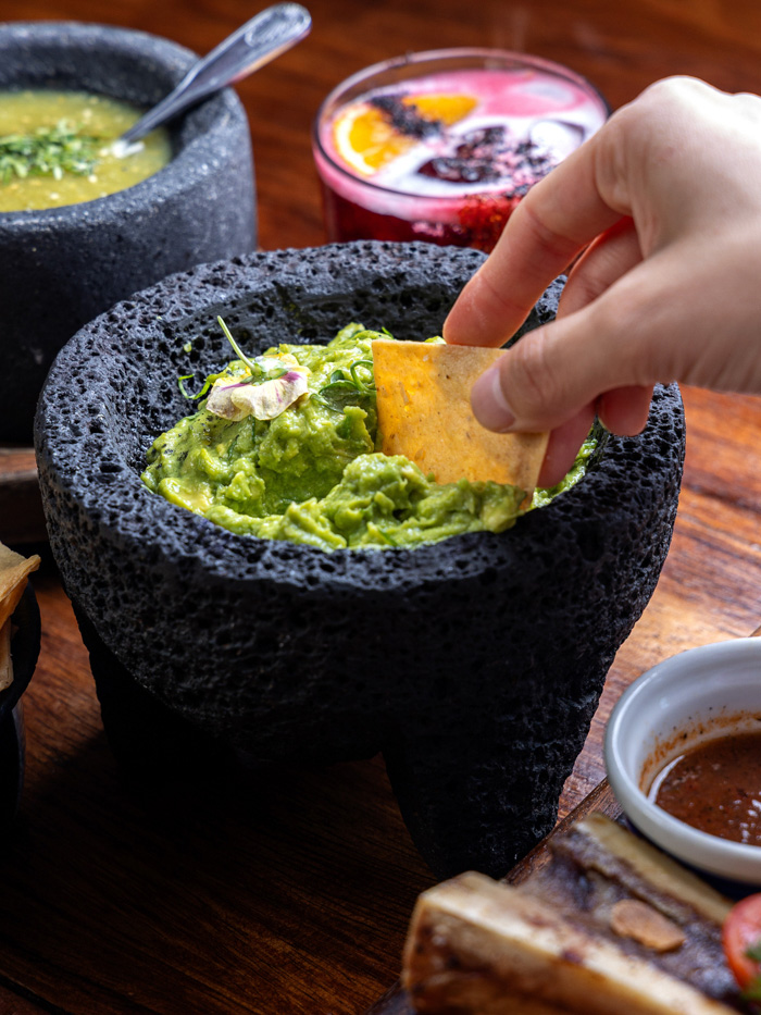 Hand dipping tortilla chip into guacamole in a molcajete, vibrant salsa and drink nearby, fun facts about Mexico