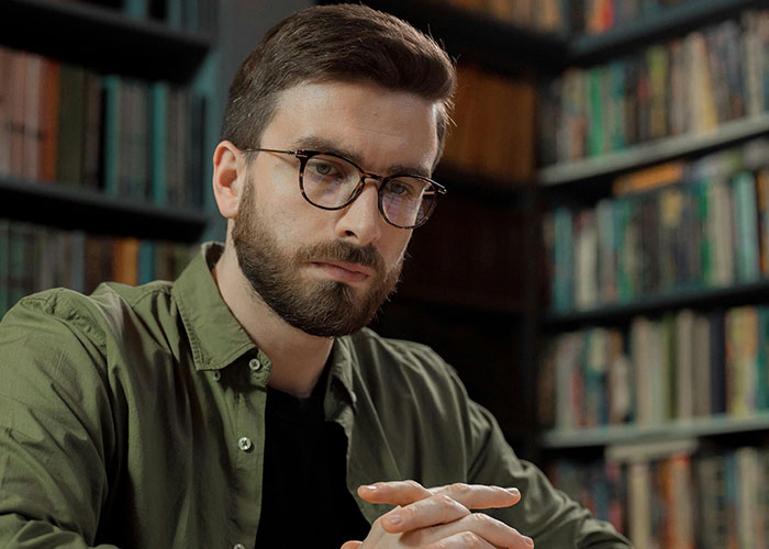 Man with glasses and beard sitting thoughtfully in a library, reflecting on life changes after learning he is not the biological parent.