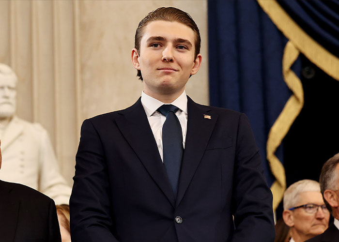 Barron Trump dressed in a suit and tie attending a formal event with a serious expression and American flag pin.