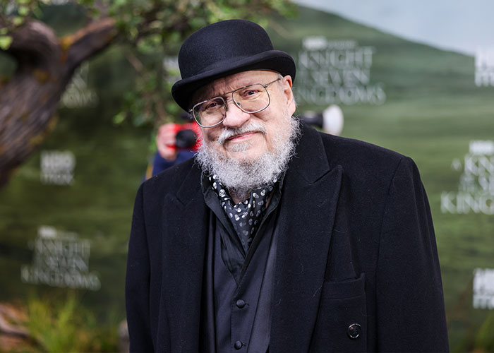 George RR Martin wearing a black coat and hat at a Knight of the Seven Kingdoms promotional event outdoors.