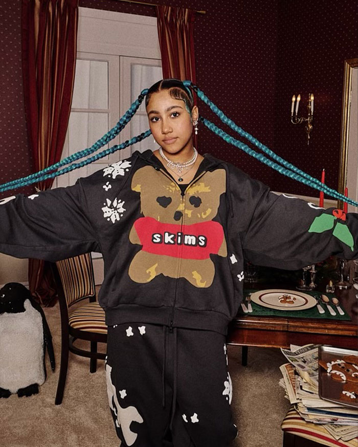 Young woman with long blue braids wearing a Skims outfit in a vintage-style room capturing a viral North West sad expression moment.