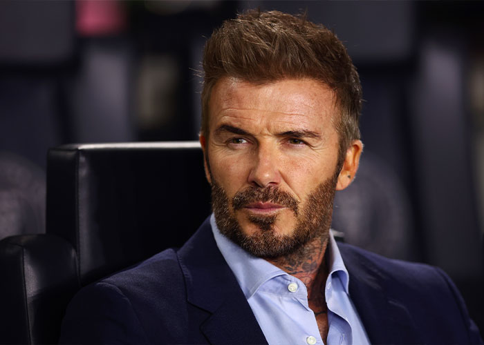 David Beckham in a dark suit and light blue shirt, seated and looking thoughtful during a public event.