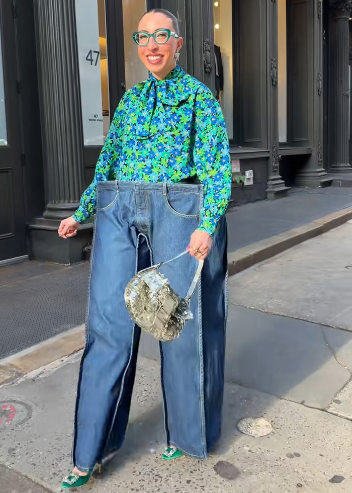 Influencer wearing oversized cubic jeans with bright floral top, green glasses, and silver handbag on a city sidewalk.