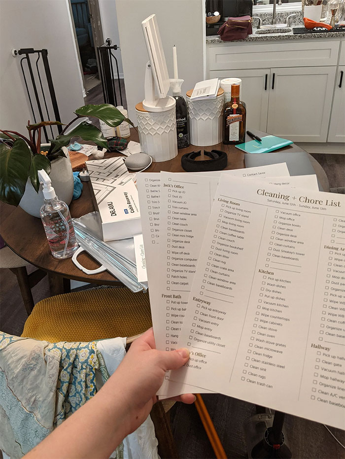 Hand holding a printed cleaning and chore list in a cluttered kitchen with household items and plants nearby.