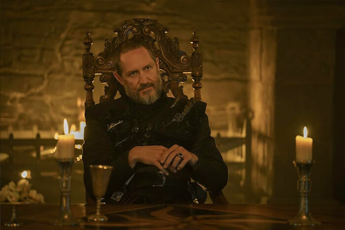 Male cast member in medieval attire sitting on an ornate throne with candles nearby from A Knight of the Seven Kingdoms season 1. Male cast member in medieval attire sitting on an ornate throne with candles nearby from A Knight of the Seven Kingdoms season 1.