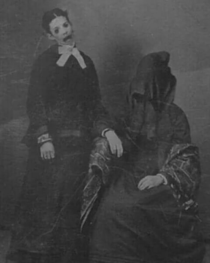 Old black and white creepy photo of two eerie figures, one with a distorted face and the other wearing a dark veil.