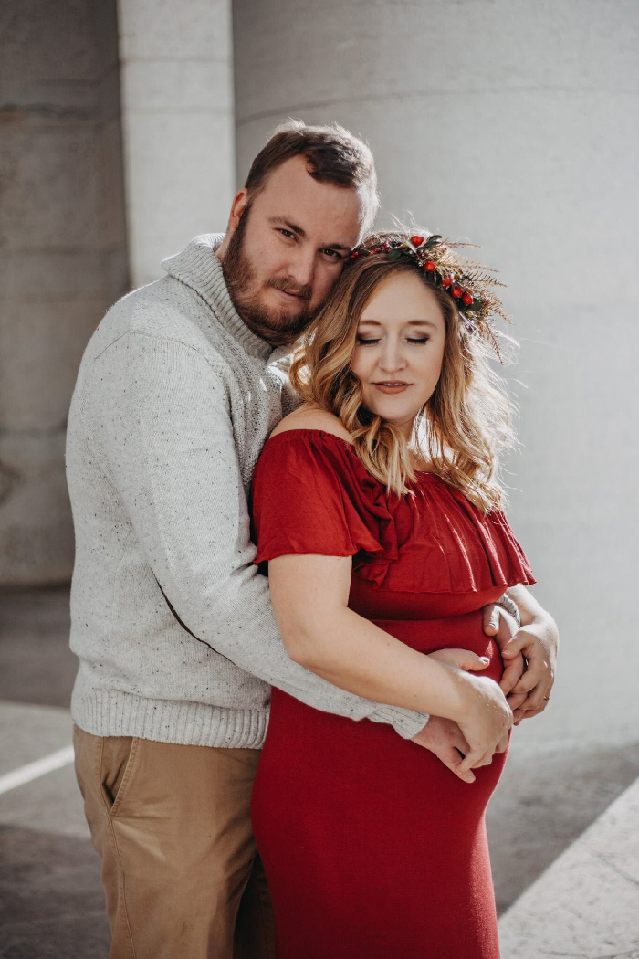Couple embracing during maternity photoshoot, partner hugging pregnant woman in red dress with floral crown